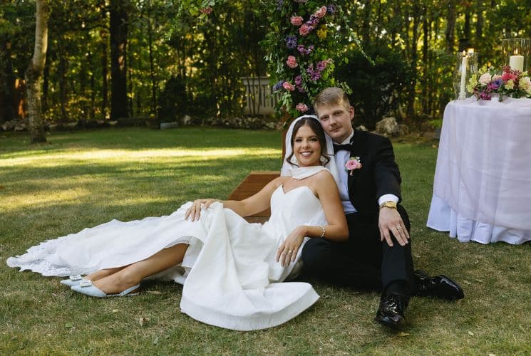 Woman wearing white dress sitting on green grass leaning on man with black suit