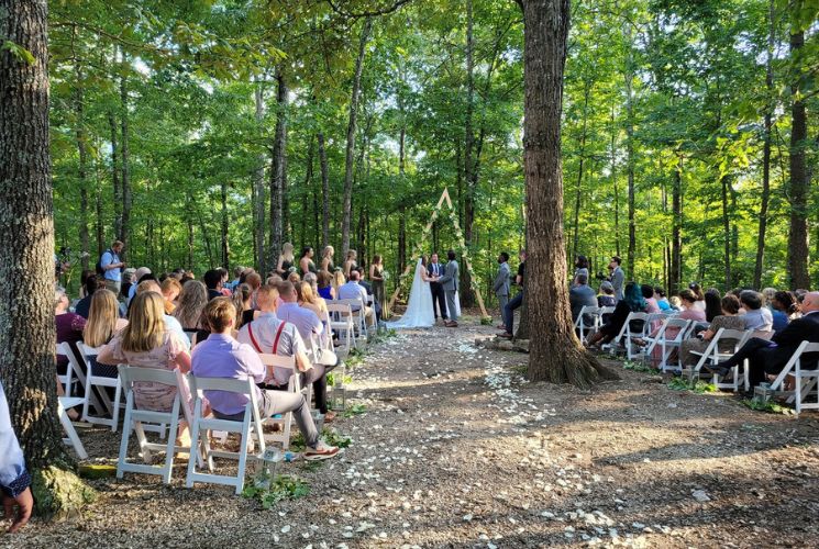 A group of people sitting in white chairs, gathered in the woods for an outdoor wedding